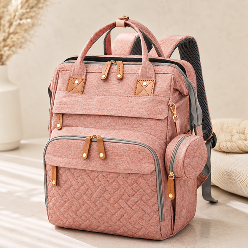 A pink textured fabric diaper backpack with tan leather-look trim and gold-tone hardware, shown standing on a light floor in a softly styled indoor setting.