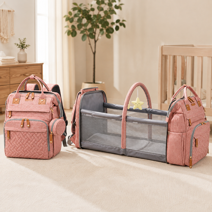 A coordinated baby travel and diaper bag set in dusty pink and gray, displayed in a softly lit nursery. The set includes a quilted diaper bag with gold-tone hardware, a matching mesh diaper bag, and a convertible bassinet with a pink frame and star mobile.