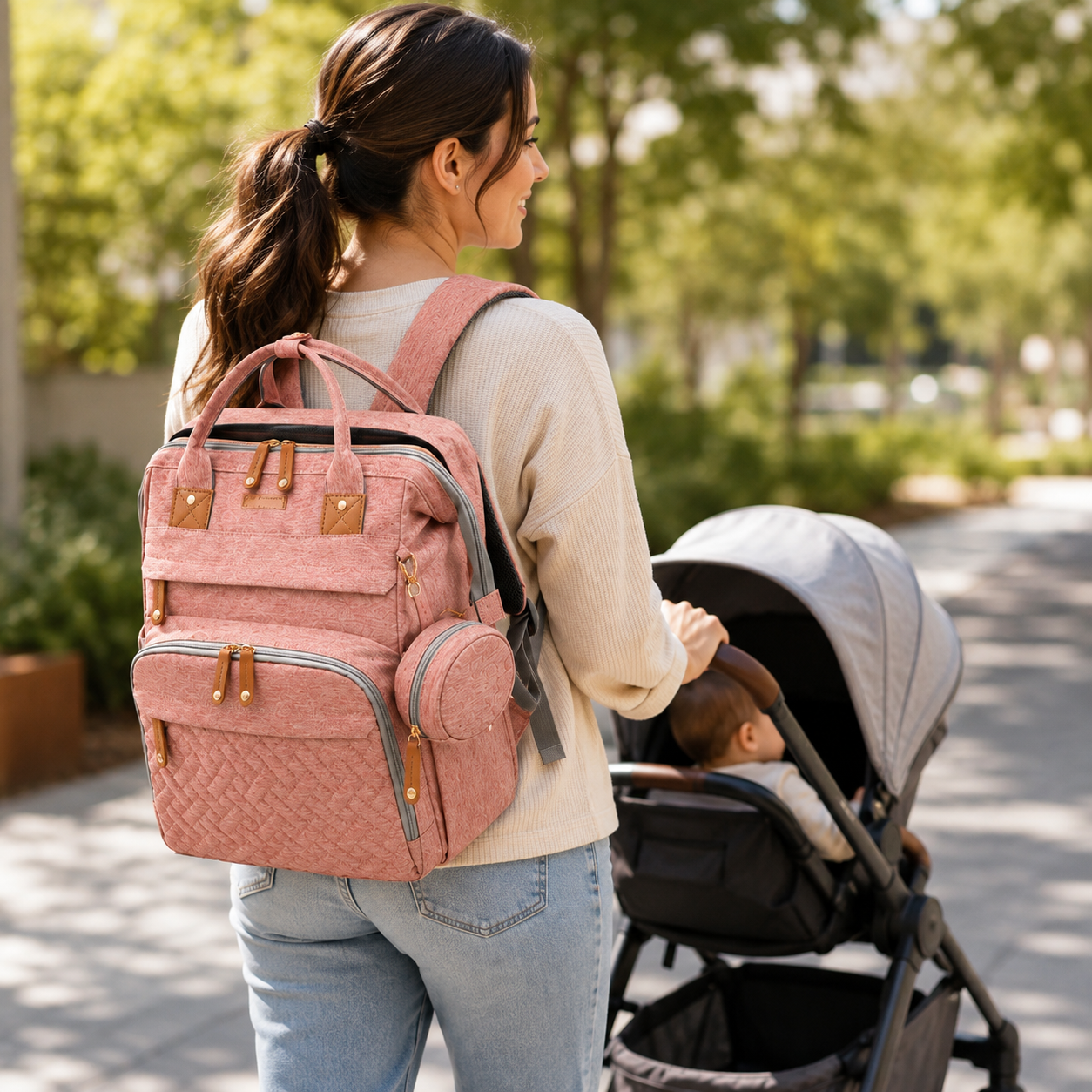 A woman wearing a pink quilted diaper backpack walks alongside a baby stroller in a sunny, tree-lined outdoor path. The backpack features multiple zippered compartments, top carry handles, and tan leather-like accents.