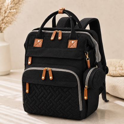 A black multi-compartment backpack with tan faux-leather accents and gray piping, shown on a neutral beige surface with a soft, styled background.