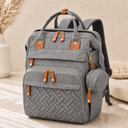 A gray fabric backpack with brown faux-leather trim and gold-tone hardware, shown standing on a light floor in a softly styled indoor setting. The bag has top handles, multiple front pockets, a side mesh pocket, and padded shoulder straps.